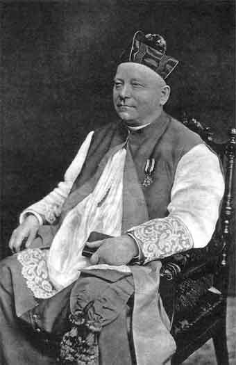 czarno-białe zdjęcie przedstawiające ks. dra Józefa Surzyńskiego siedzącego na rzeźbionym krześle, ubranego w strój duchownego, w czapce na głowie i medalem na piersi